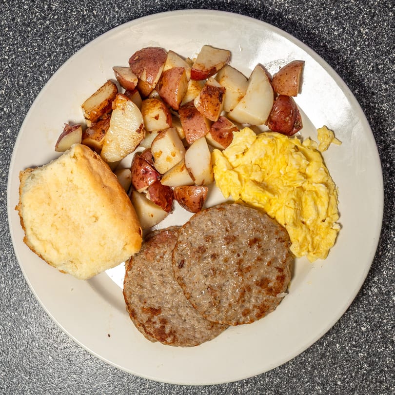 A diner breakfast plate with scrambled eggs, sausage patties, home fries, and a biscuit.
