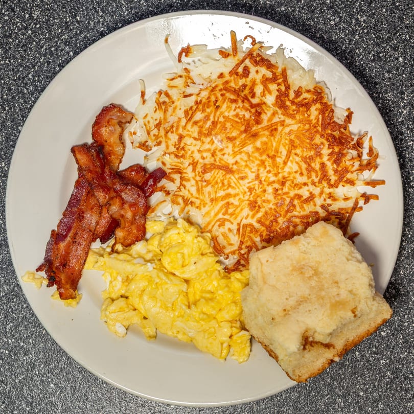 A diner breakfast plate with crispy bacon, scrambled eggs, golden hash browns, and a biscuit.